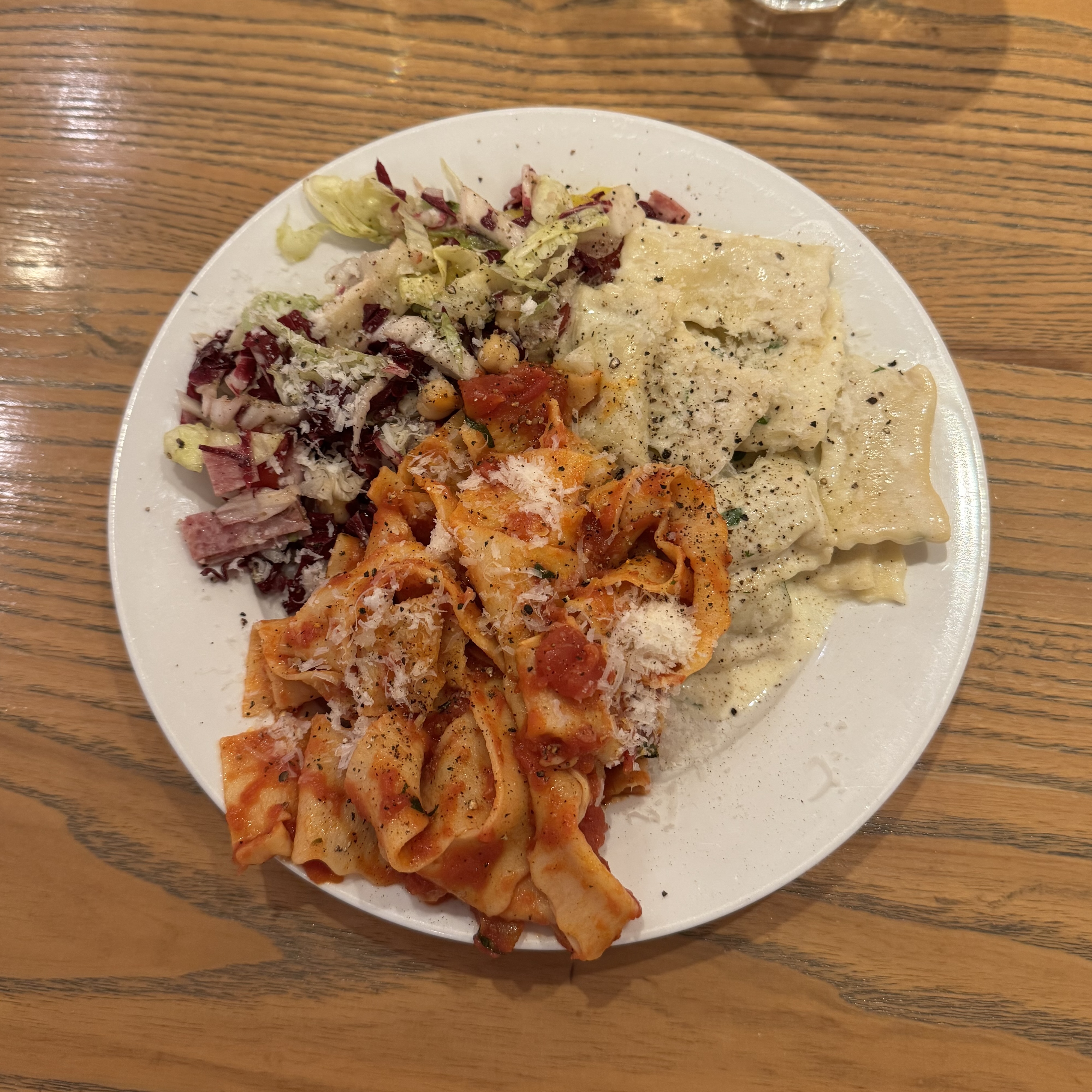 A white plate on a wooden table holds three pasta dishes: ribbon pasta with tomato sauce and grated cheese, plain ravioli with black pepper, and a chicory salad with shaved parmesan. Each portion is arranged in its own section on the plate, creating a trio sampler.