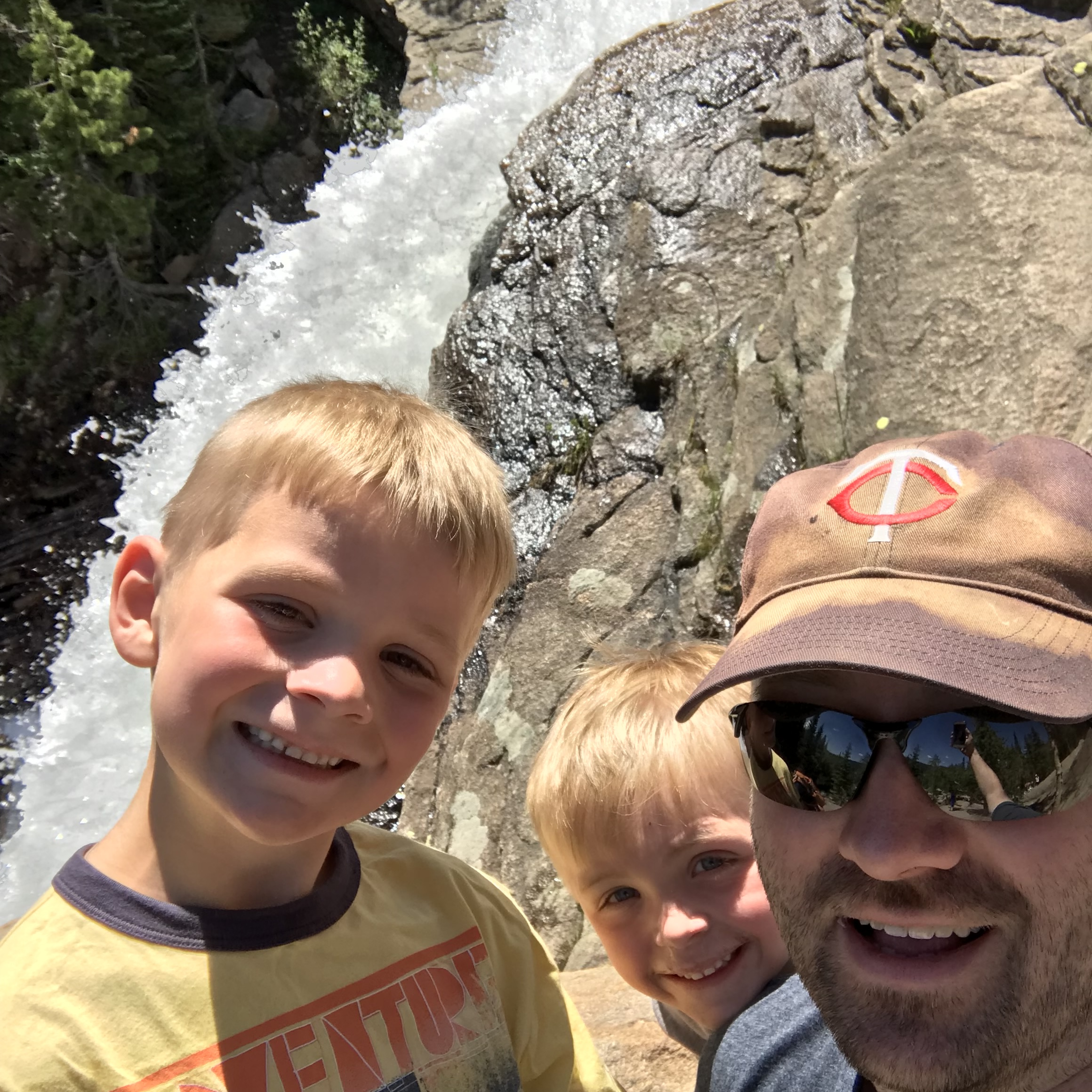 A smiling man in sunglasses and a baseball cap takes a selfie with two young blonde boys at a rushing waterfall. White water cascades over large gray rocks in the background, surrounded by green foliage. All three are grinning at the camera on a sunny day outdoors.