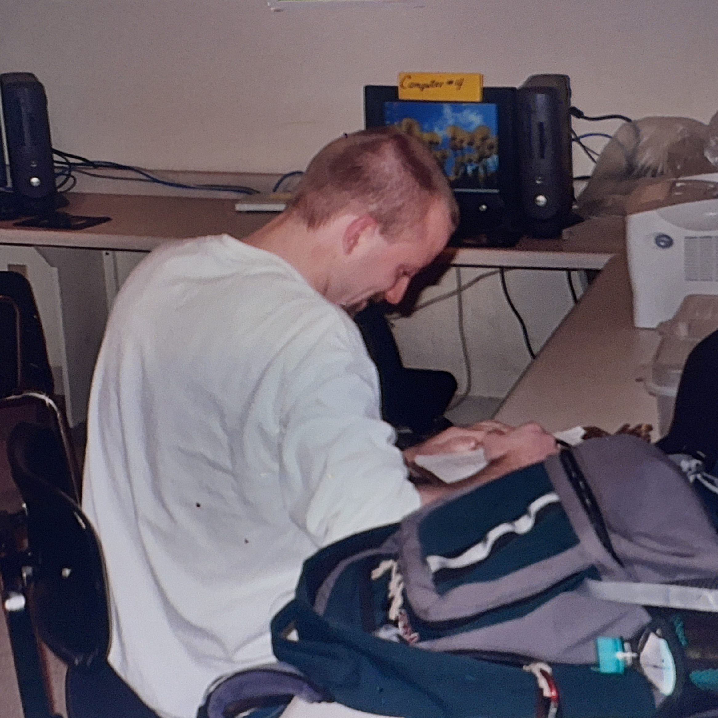 A young person in a white long-sleeve shirt sits at a home computer desk, focused on reading or writing in a notebook. The desktop setup includes a CRT monitor, speakers, and various cables. A backpack and school supplies are visible nearby, suggesting homework or studying time.