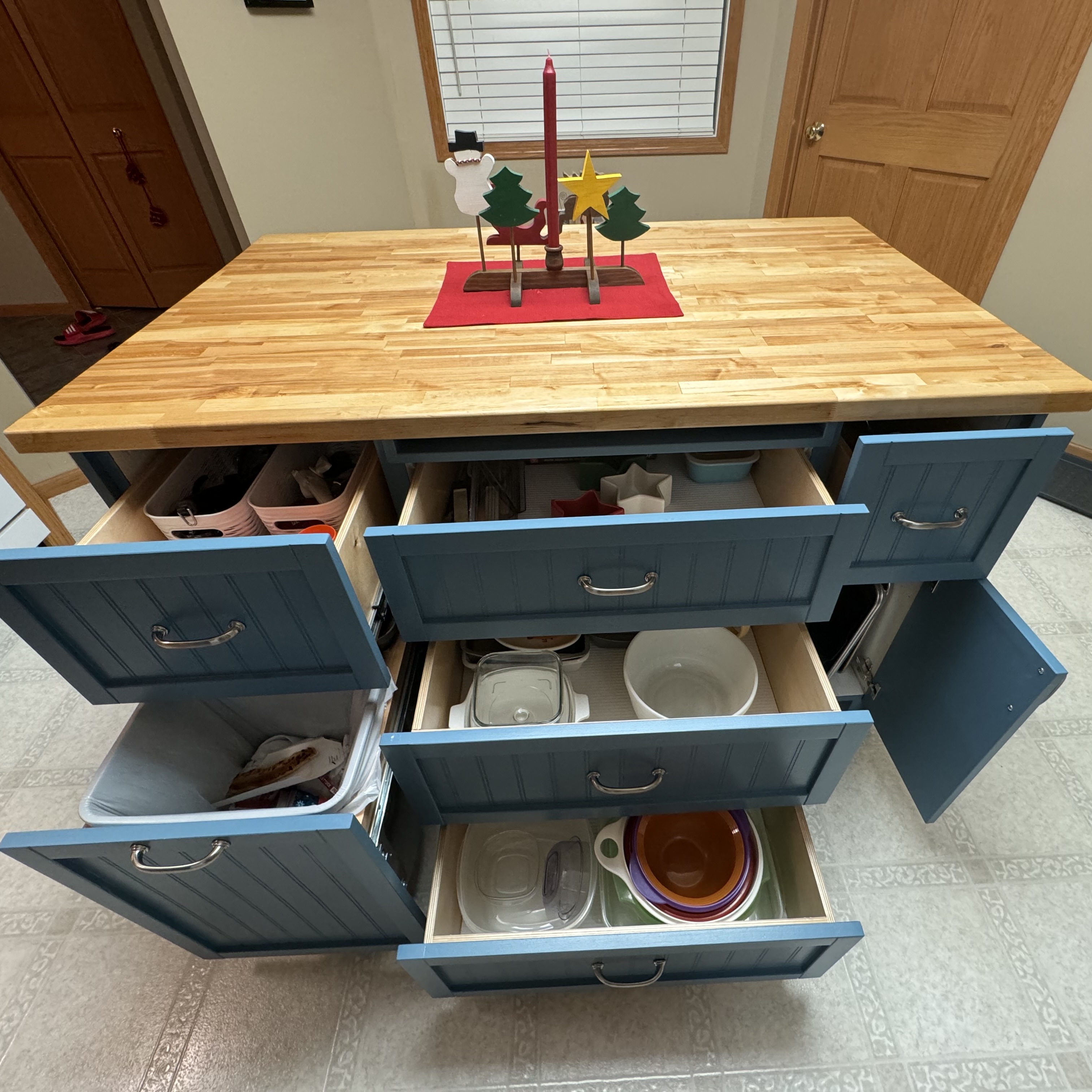 A kitchen island with butcher block top and teal-painted drawers, all fully opened to reveal organized storage of dishes, food containers, and kitchen items. A simple holiday centerpiece with a candle, star, and Christmas tree decorations sits on a red placemat on top.