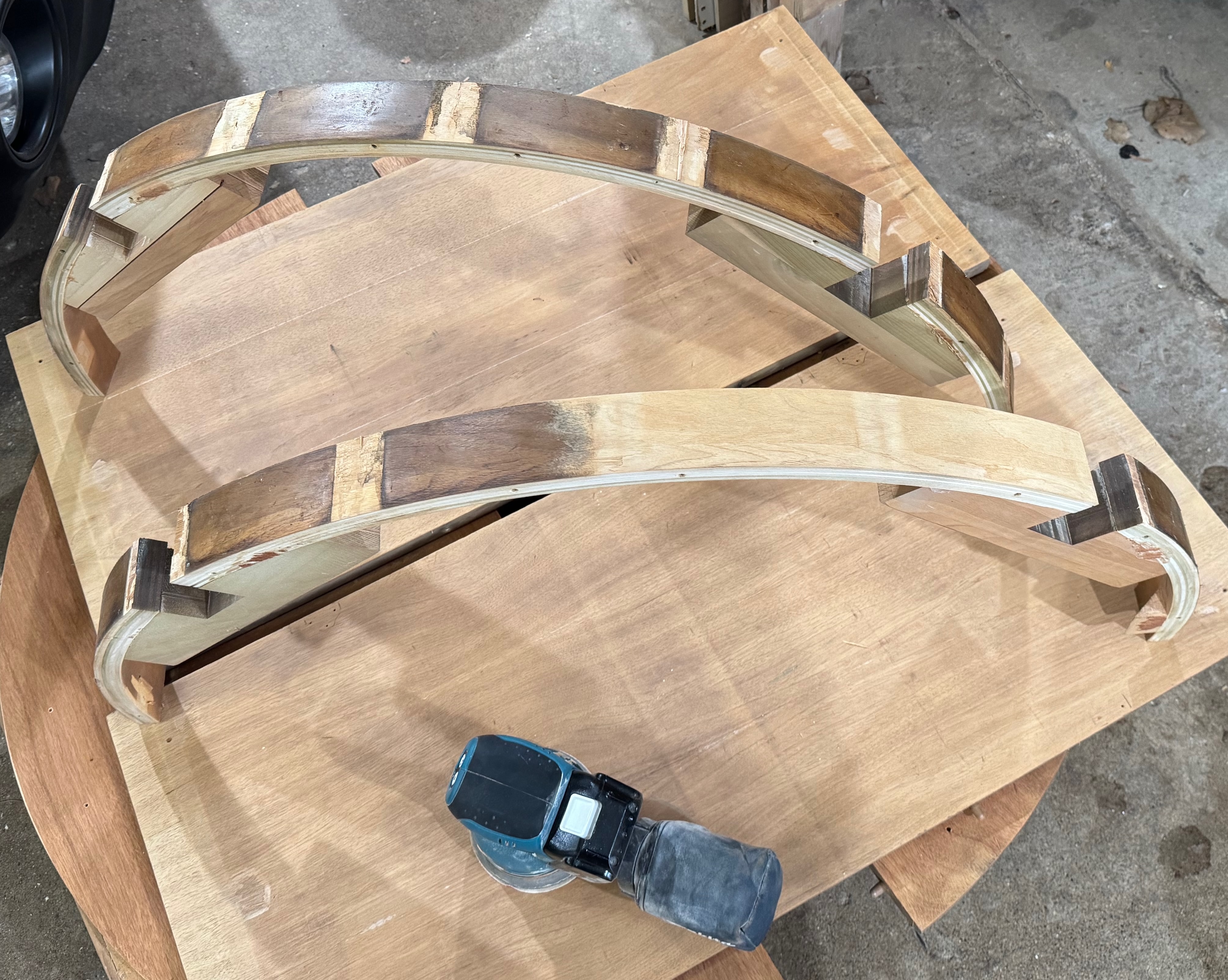 Two curved wooden pieces rest on a workbench beside a sander, indicating ongoing woodworking.